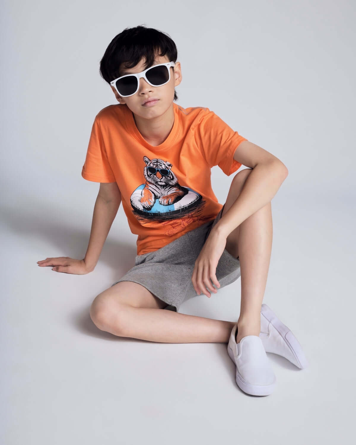 Child wearing an orange t-shirt with a tiger graphic, gray shorts, and white sneakers on a light gray background.
