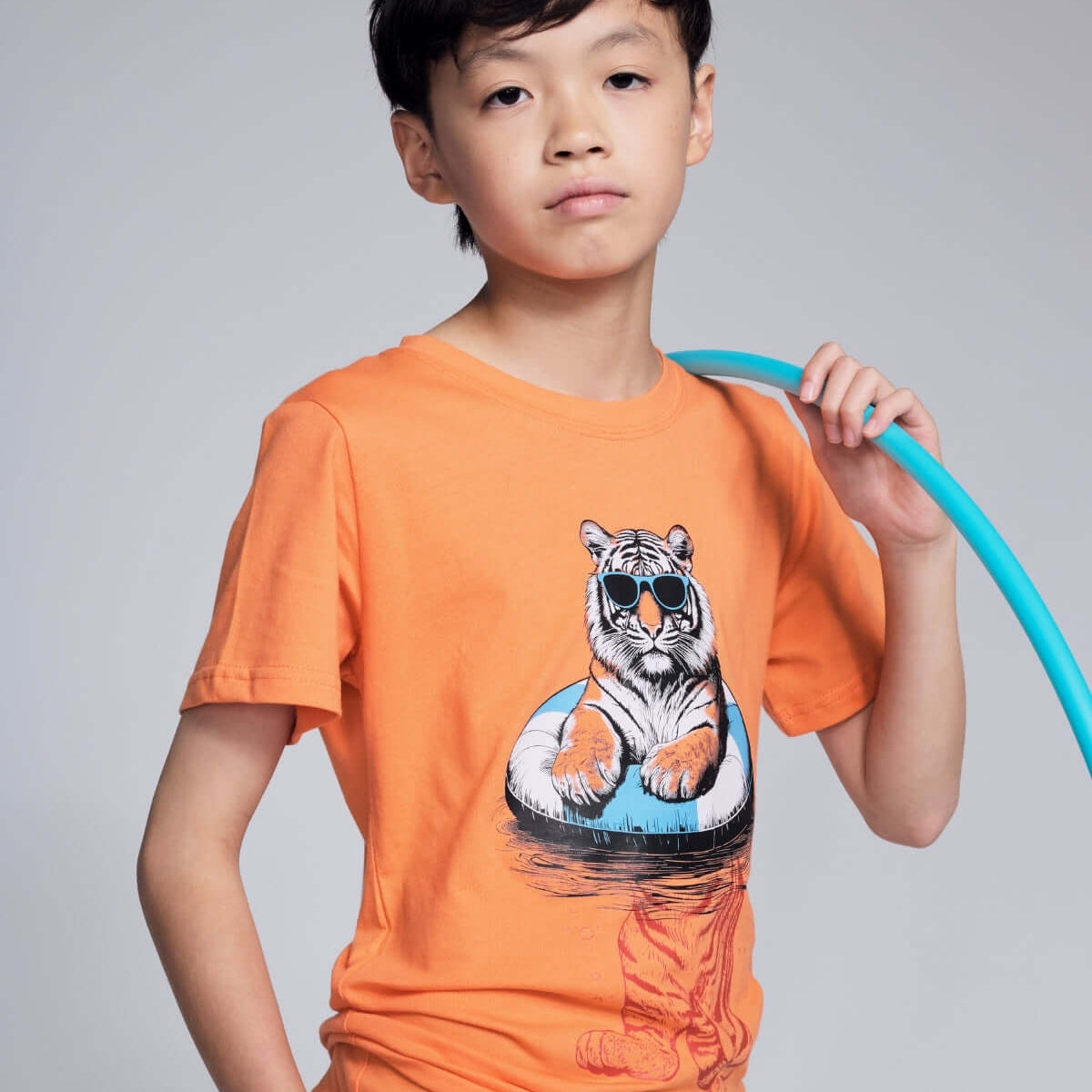 Child wearing an orange t-shirt with a tiger graphic, holding a blue hula hoop against a gray background