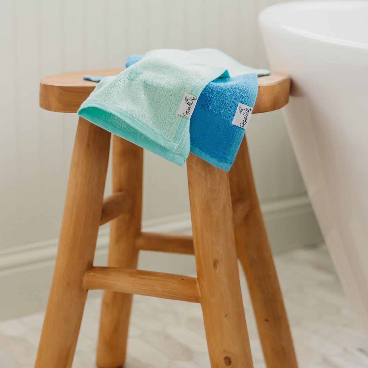 Two towels, one green and one blue, draped over a wooden stool in a bathroom setting.