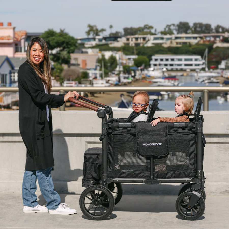 Woman pushing a black WonderFold stroller with two children on a waterfront boardwalk.