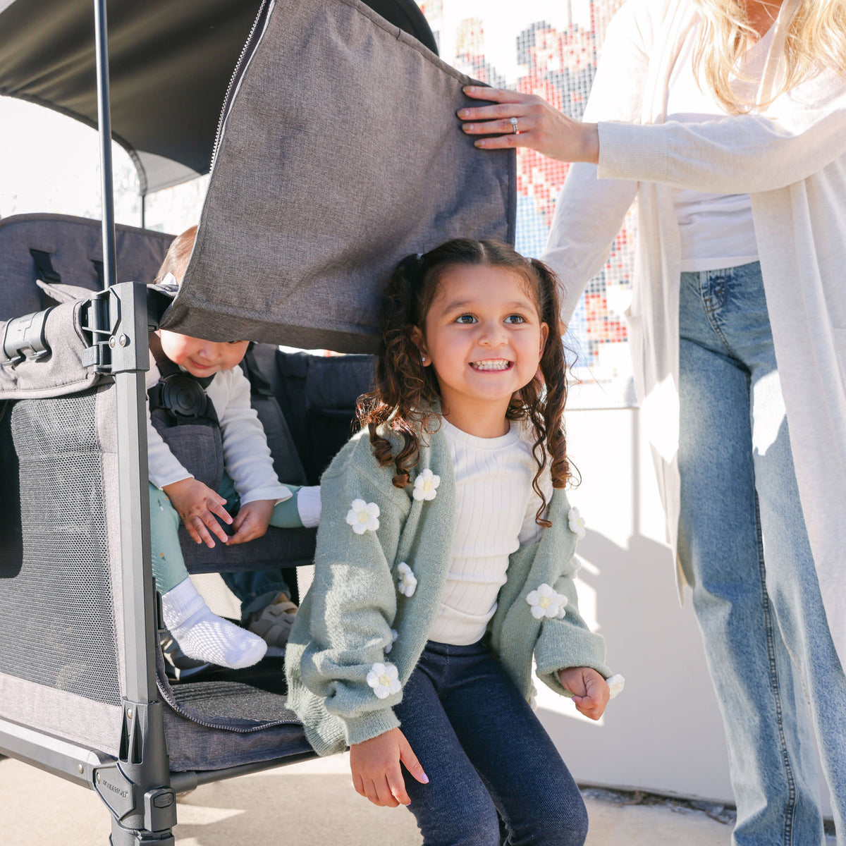 Child in a green coat standing next to a stroller with a person in the background.
