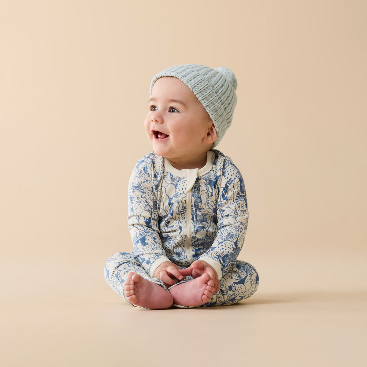 Baby wearing a patterned onesie and knitted hat sitting on a beige background