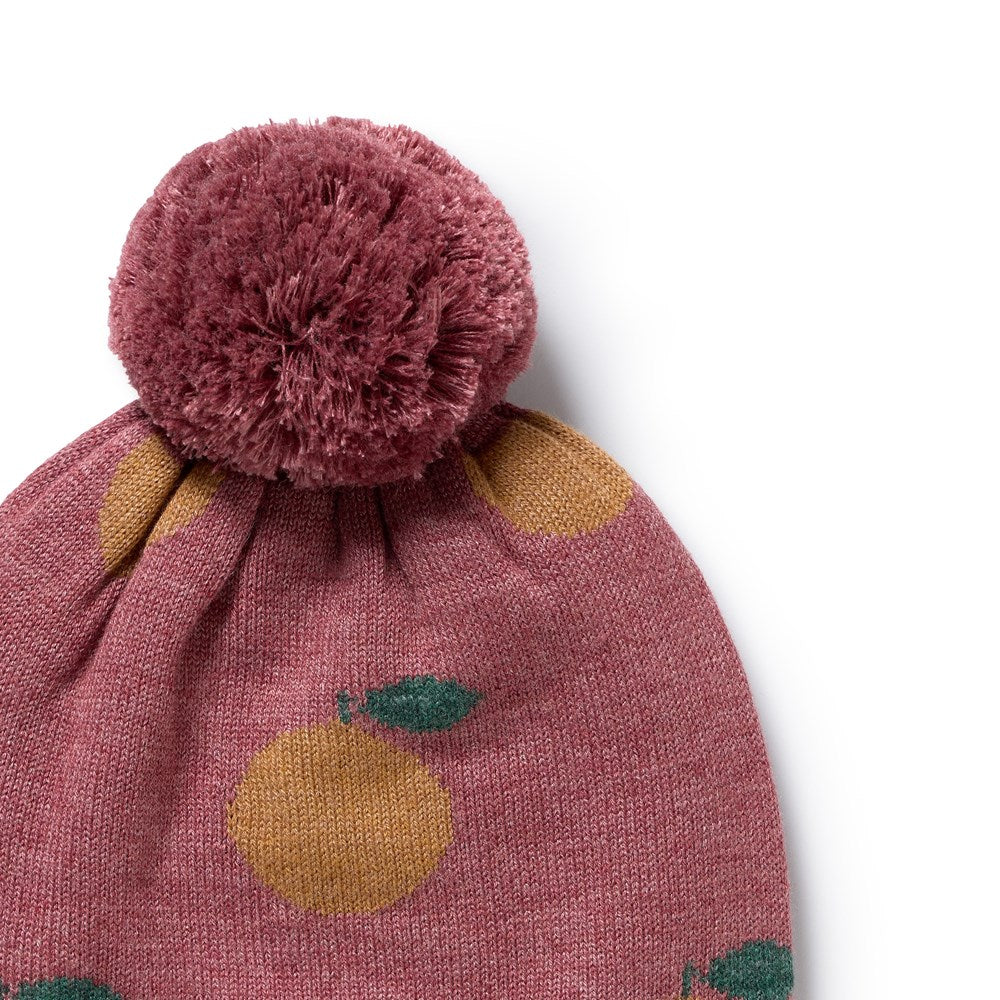 A close-up of a knit pom-pom beanie in dusty rose pink with a yellow and green fruit pattern, featuring a large textured pom-pom on top, set against a plain white background.