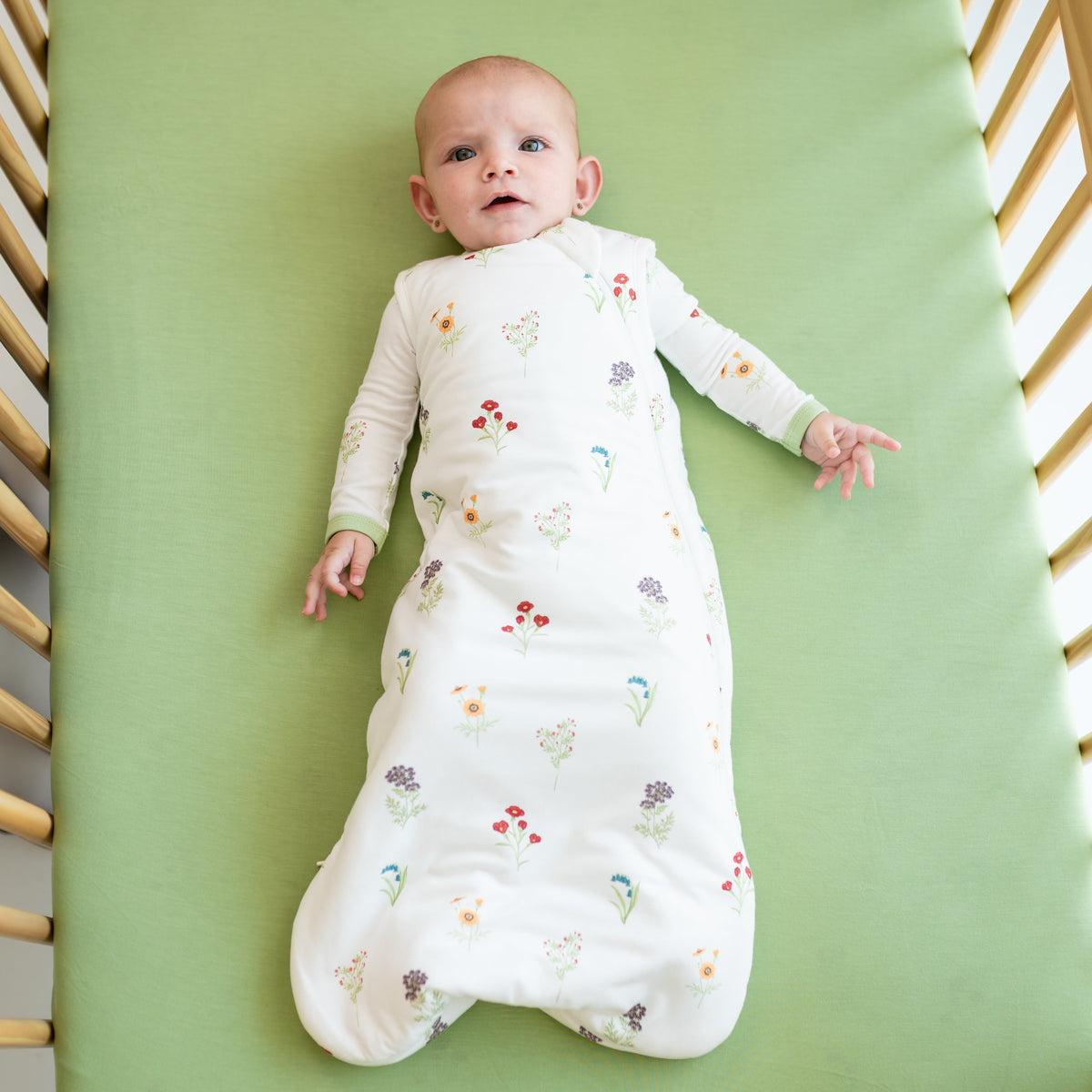 Baby lying in a crib wearing a floral-patterned onesie on a green mattress.
