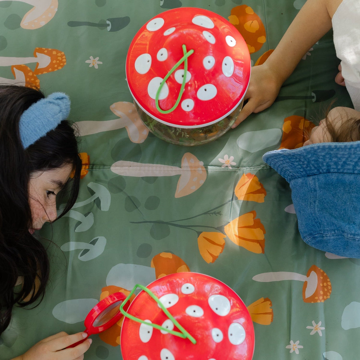 Two children playing with red paper clocks on a green tablecloth with mushroom patterns.