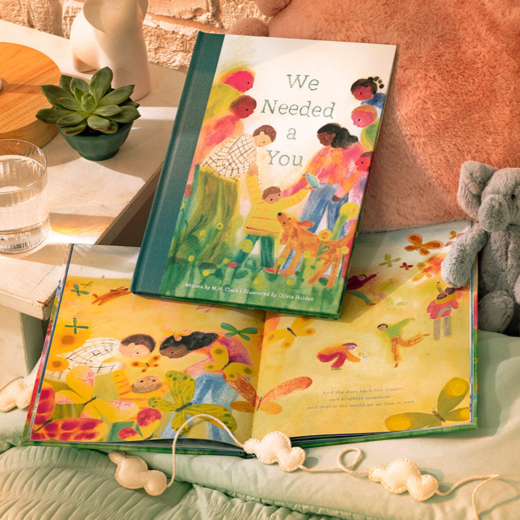 Children's book titled 'We Needed a You' on a bed with a teddy bear and plant in the background.