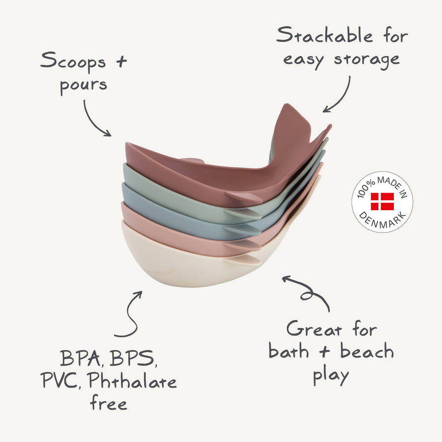 A stack of five whale-shaped bath toys with handwritten-style labels highlighting key product features: "Scoops + pours," "Stackable for easy storage," "BPA, BPS, PVC, Phthalate free," and "Great for bath + beach play," alongside a "100% Made in Denmark" circular badge.