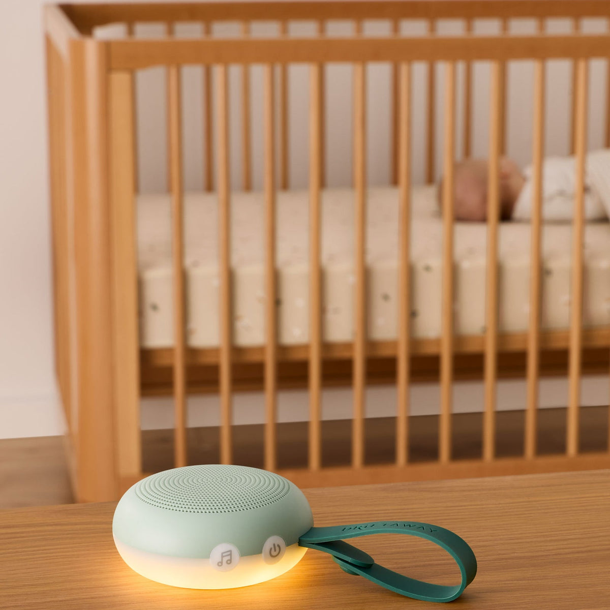 Mint green baby monitor with a glowing base on a wooden surface, with a crib and baby in the background.