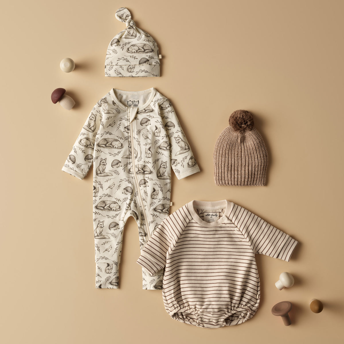 A baby clothing set arranged on a warm beige background, including a long-sleeve footless romper with an allover animal print, a matching bonnet, a ribbed knit pom-pom hat, and a long-sleeve striped bodysuit. Small mushroom props are scattered around the items.