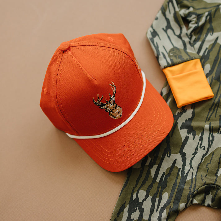 Orange cap with deer emblem on a camouflage jacket against a beige background