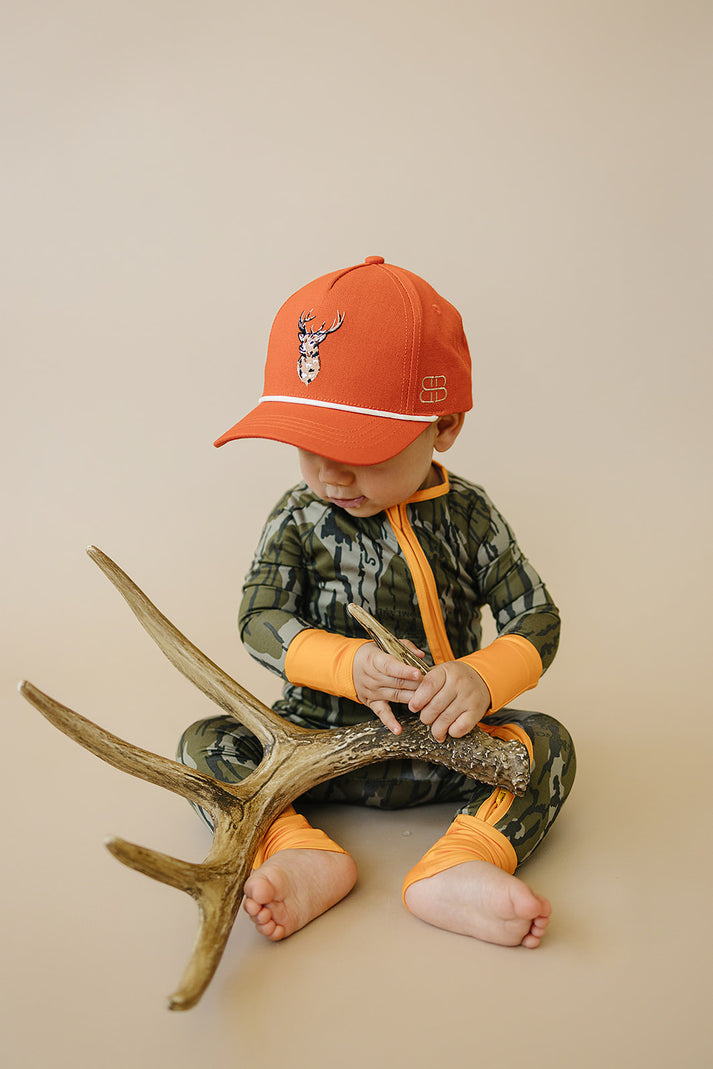 Child wearing an orange cap and camouflage outfit sitting on a large deer antler against a beige background