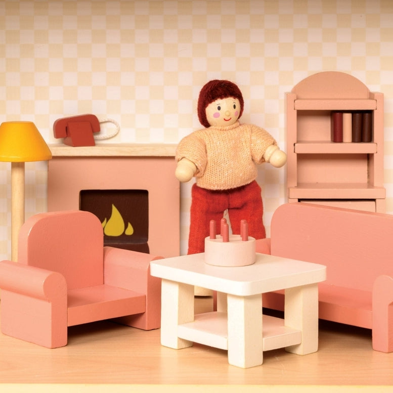 A miniature wooden dollhouse room interior featuring a wooden toy figure standing next to a coral-pink sofa and armchair set, a coffee table with a tiny red telephone, a pale pink decorative fireplace, a yellow floor lamp, and a natural wood bookcase against a patterned wallpaper background.