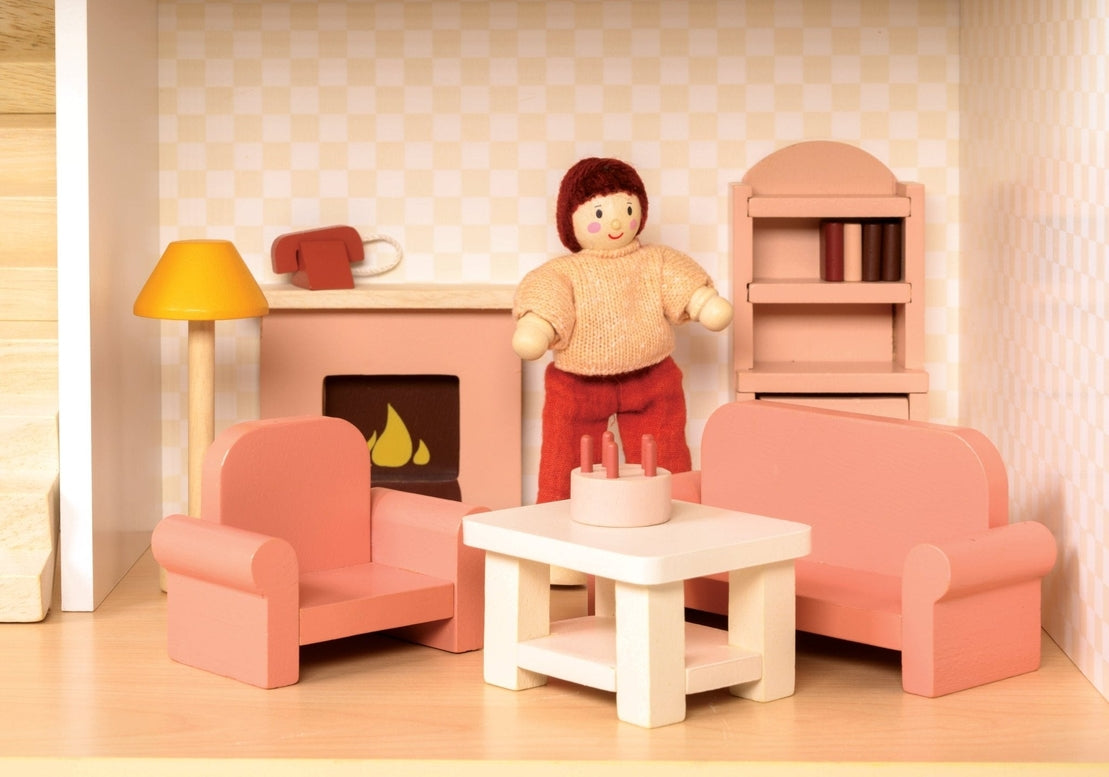 A miniature wooden dollhouse room interior featuring a wooden toy figure standing next to a coral-pink sofa and armchair set, a coffee table with a tiny red telephone, a pale pink decorative fireplace, a yellow floor lamp, and a natural wood bookcase against a patterned wallpaper background.