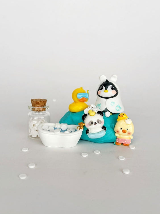 A small collection of cute, colorful polymer clay figurines, including a penguin in a white swimsuit, a yellow rubber duck with goggles, a panda, a chick, and a miniature white bathtub with a cork stopper, arranged on a light gray background.