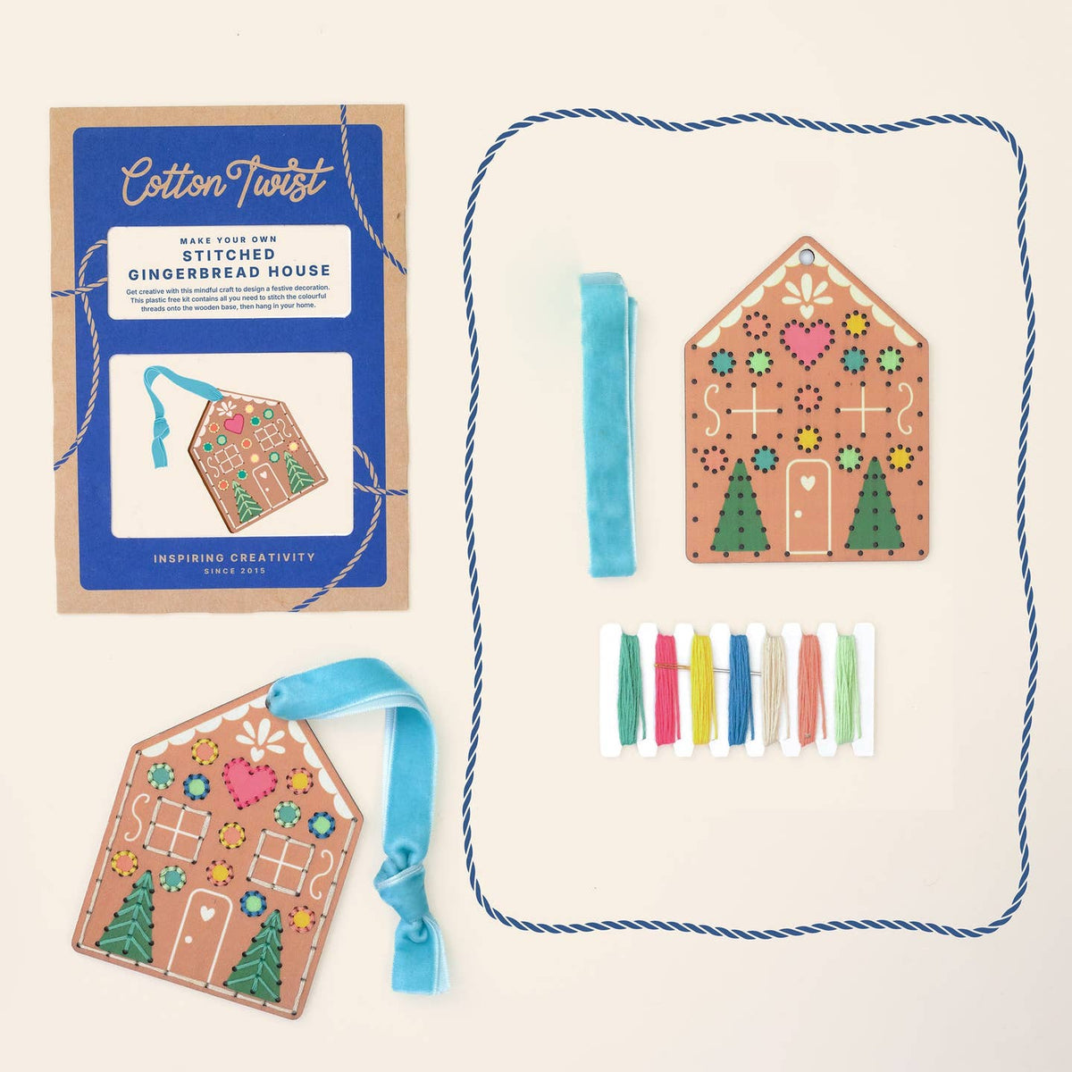 A flat-lay view of a 'Cotton Twist' gingerbread house stitching kit, showing the blue and kraft paper packaging, a completed decorated gingerbread house ornament, a blue ribbon, colorful thread spools, and a blue rope border on a light cream background.