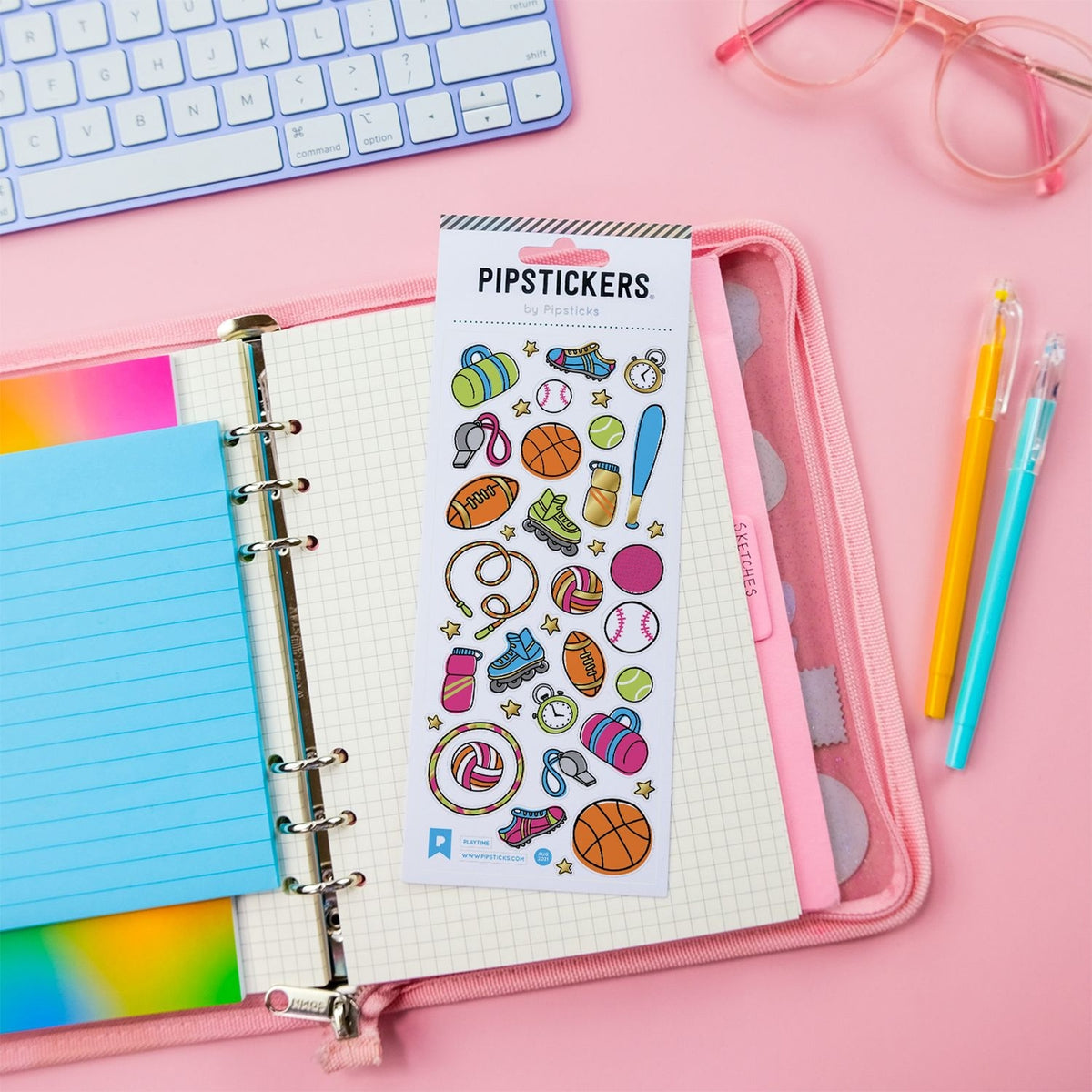 A sheet of colorful sports-themed lip stickers labeled 'PIPSTICKERS by Stephanie' placed on an open pink ring binder notebook, surrounded by a keyboard, glasses, pens, and a pastel notepad on a pink background.