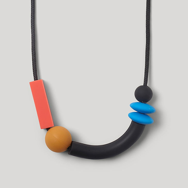 A minimalist geometric beaded necklace featuring a black cord, a large black curved bar, a round tan bead, a bright coral-red rectangular bead, a blue spiral bead, and a small black bead at the end.
