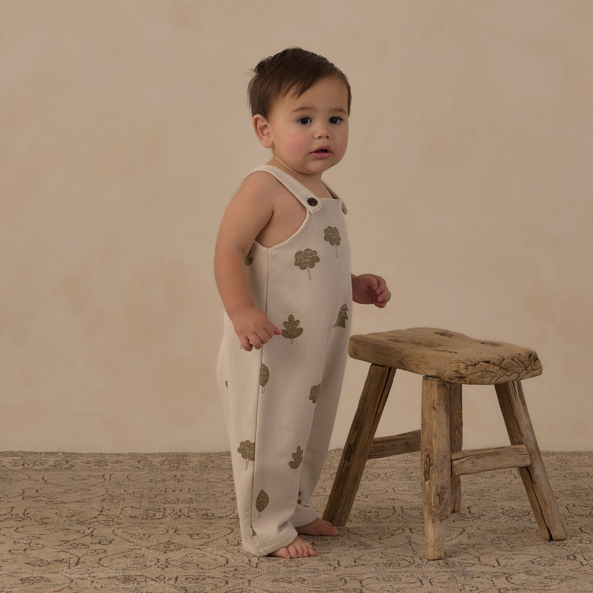 A baby standing barefoot next to a rustic wooden stool, wearing a cream-colored sleeveless jumpsuit with a brown tree-leaf pattern, set against a warm beige textured backdrop.