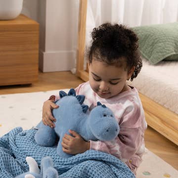 A young child sitting on a floor in a bedroom, holding a light blue plush dinosaur toy under a blue blanket.