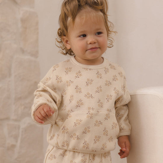 A toddler with curly light brown hair wearing a cream-colored waffle-knit long-sleeve top and matching pants with a small floral pattern, standing indoors next to a white chair.