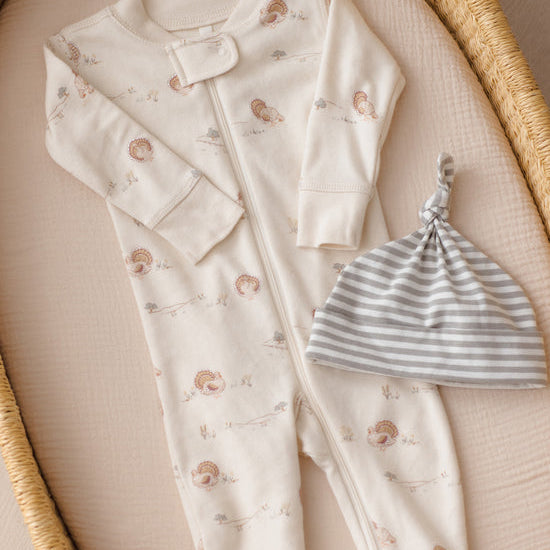 A baby one-piece footless sleeper in cream with a small hedgehog print, laid flat on a beige cushion inside a woven tray, next to a gray and white striped knit beanie.