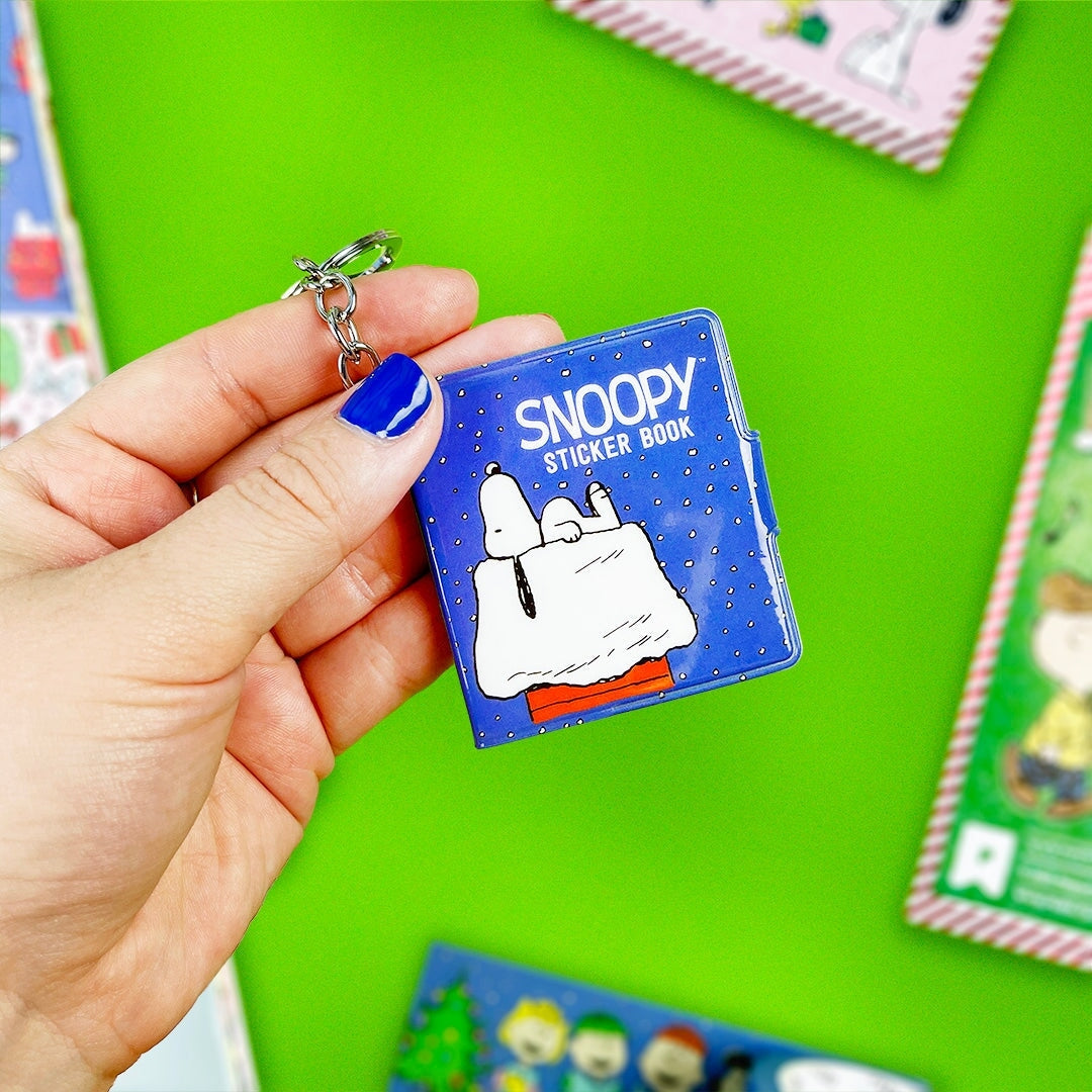 Hand holding a Snoopy sticker book keychain against a green background