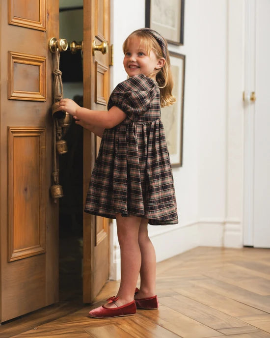 A young girl with blonde hair stands in a doorway wearing a brown and black plaid dress and red shoes, holding the door open with one hand.