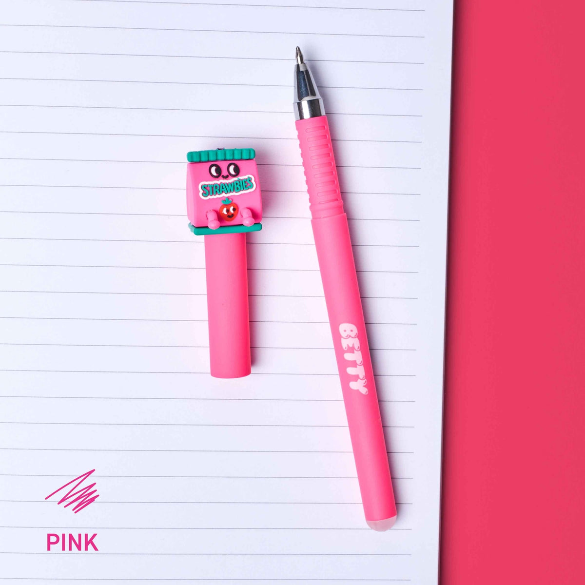 A pink retractable pen and matching pink highlighter with a cartoon character charm labeled 'STROKES' are arranged on a lined notebook page next to a solid pink background. The word 'PINK' appears in the bottom-left corner.