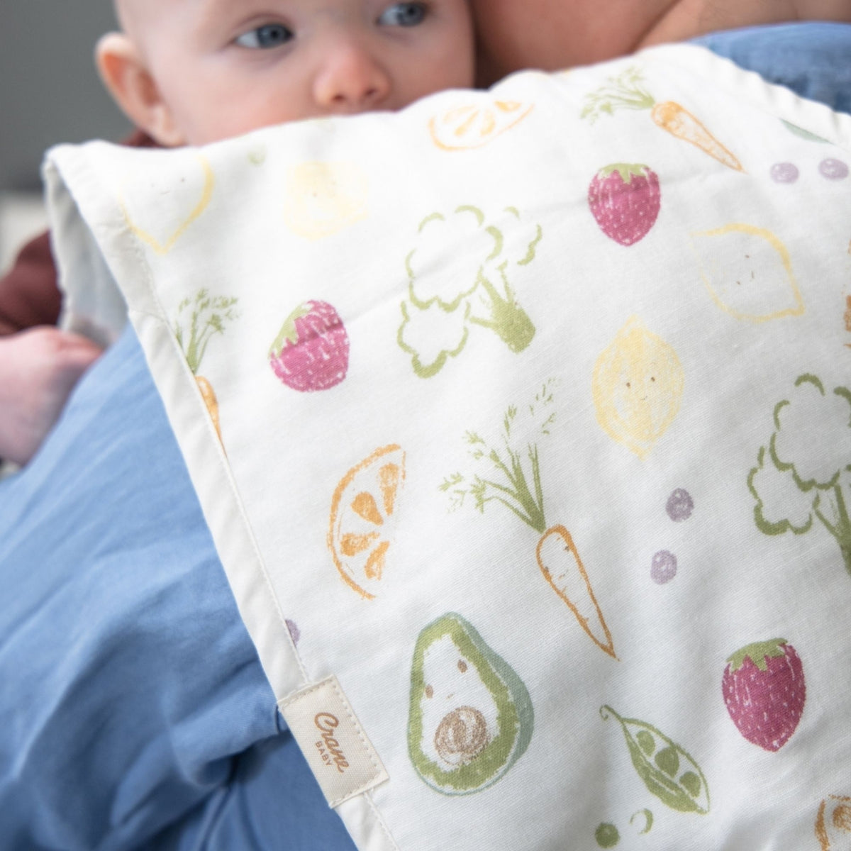 Baby wrapped in a blanket with fruit and vegetable patterns, held by an adult.