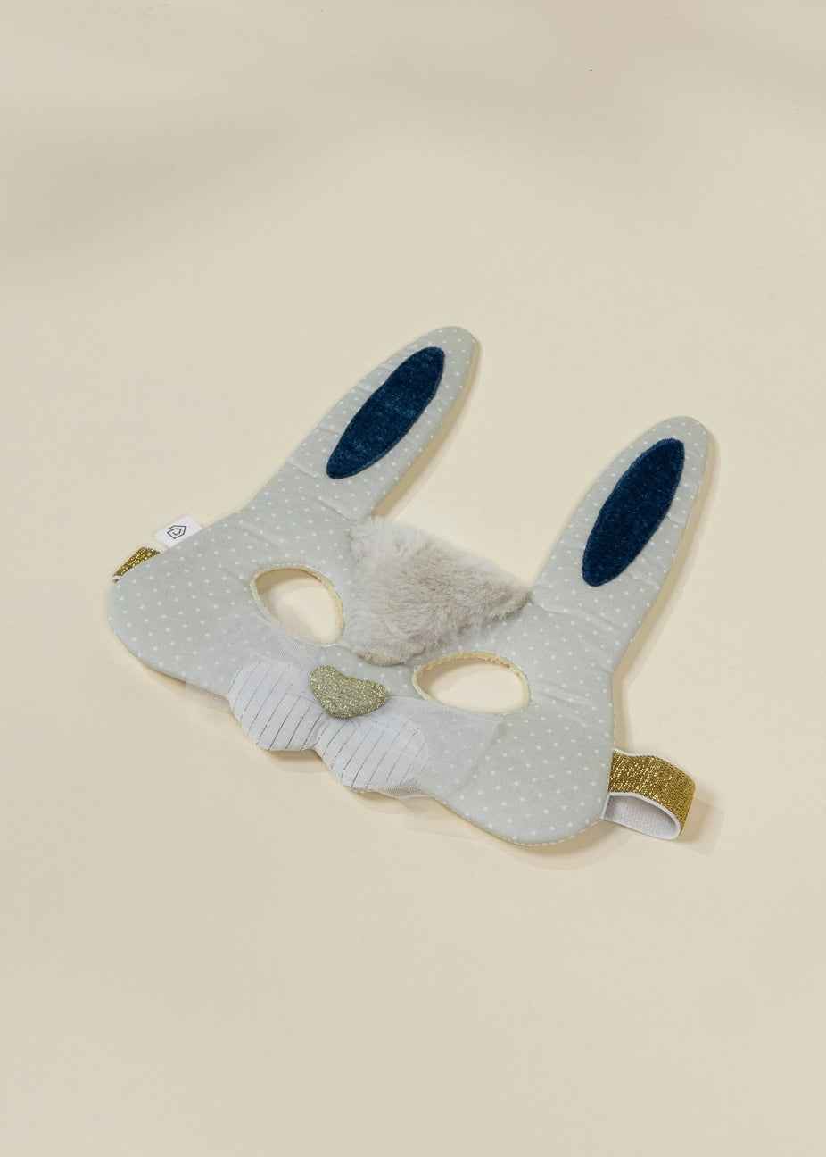 A white fabric bunny mask with dotted pattern, featuring blue inner ears, a fluffy white faux fur nose, and gold glitter trim on the ear tips and side straps, shown on a light beige background.