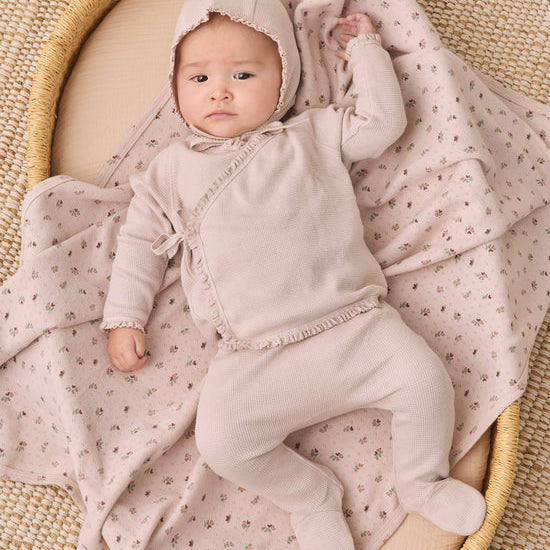 A baby wearing a light beige footed onesie and bonnet lies on a cream woven mat, wrapped in a matching beige and pink floral blanket.