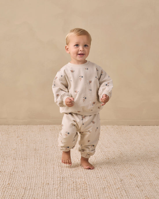 A toddler standing barefoot on a textured beige rug against a warm beige wall, wearing a cream-colored long-sleeve top and matching pants with a small scattered animal print.