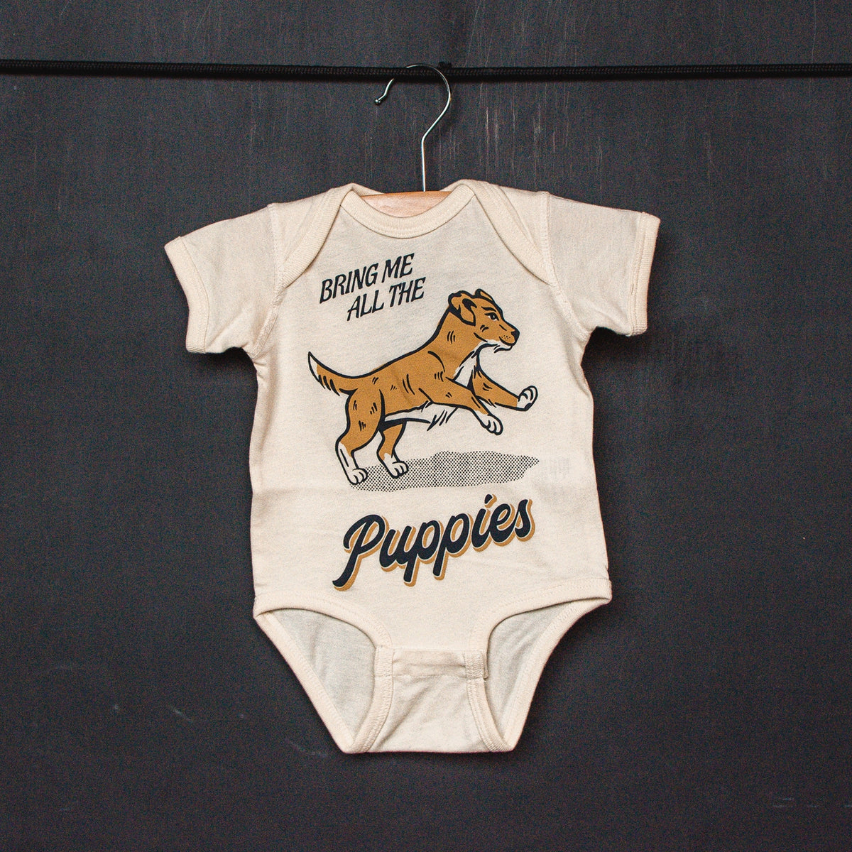 An off-white short-sleeve baby bodysuit laid flat on a textured surface, featuring a graphic of a yellow and white puppy with the text 'BRING ME ALL THE Puppies'.