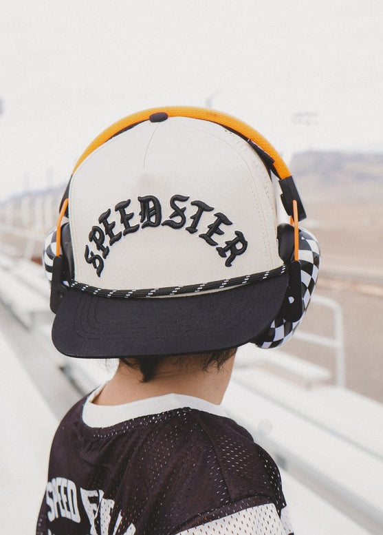 A person viewed from behind wearing a cream and black baseball cap with 'SPEEDSTER' embroidered in black across the front, along with orange headphones and a dark mesh jersey in an outdoor setting.