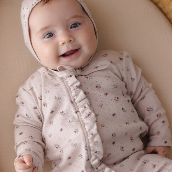 A smiling baby lying on a beige cushion, wearing a light pink knit romper and bonnet with a small floral print.