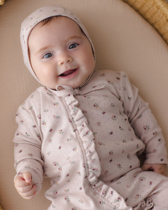A smiling baby lying on a beige cushion, wearing a light pink knit romper and bonnet with a small floral print.