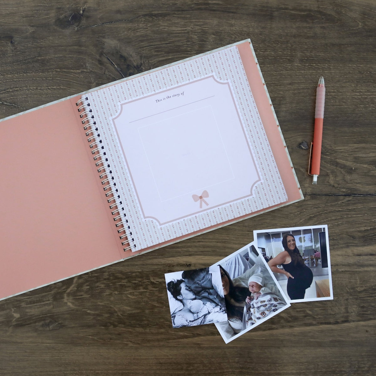 Open spiral-bound notebook with pink cover and white pages on a wooden surface, accompanied by a pen and two photos.