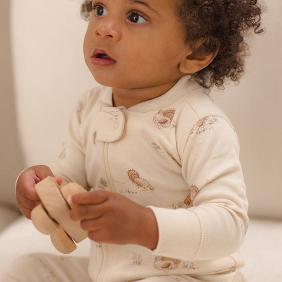 A baby wearing a cream-colored long-sleeve footless onesie with small brown animal illustrations, holding a wooden toy while looking off-camera.