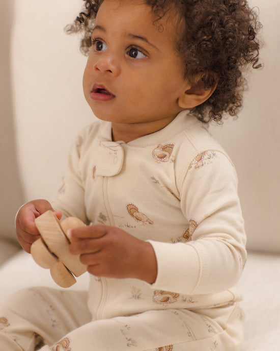 A baby wearing a cream-colored long-sleeve footless onesie with small brown animal illustrations, holding a wooden toy while looking off-camera.