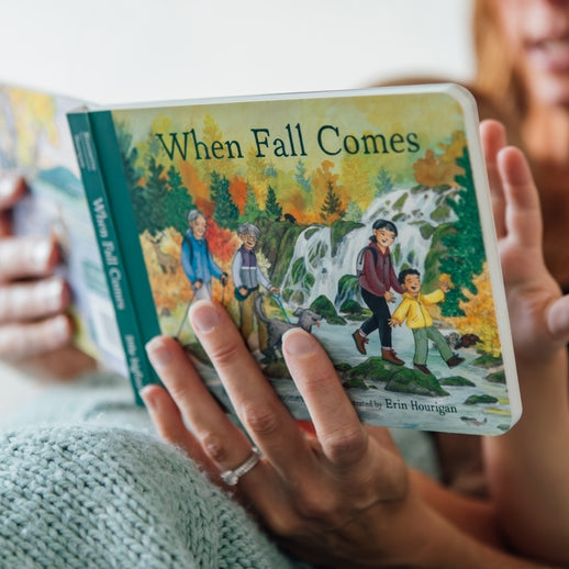 A toddler holding an open board book titled When Fall Comes, looking down at the colorful illustrated pages showing autumn trees and an outdoor nature scene.