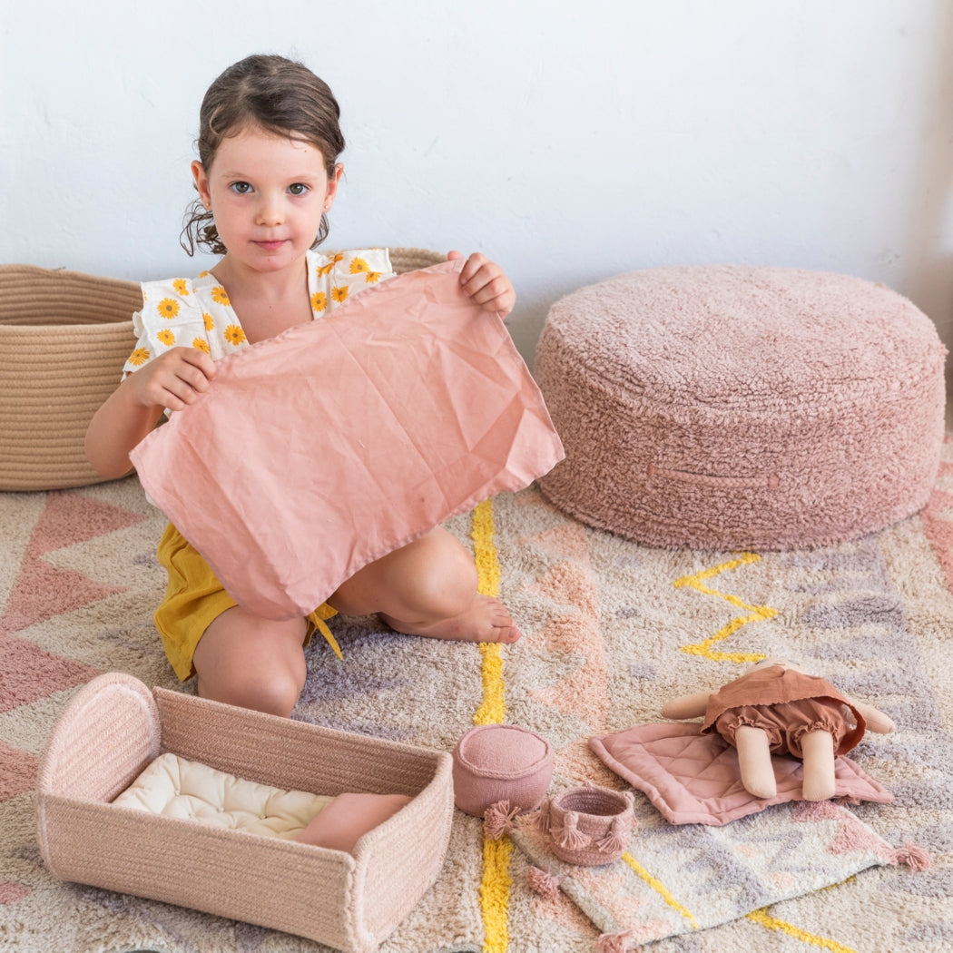 Young girl holding a pink plush toy in a room with soft furnishings and toys on the floor.