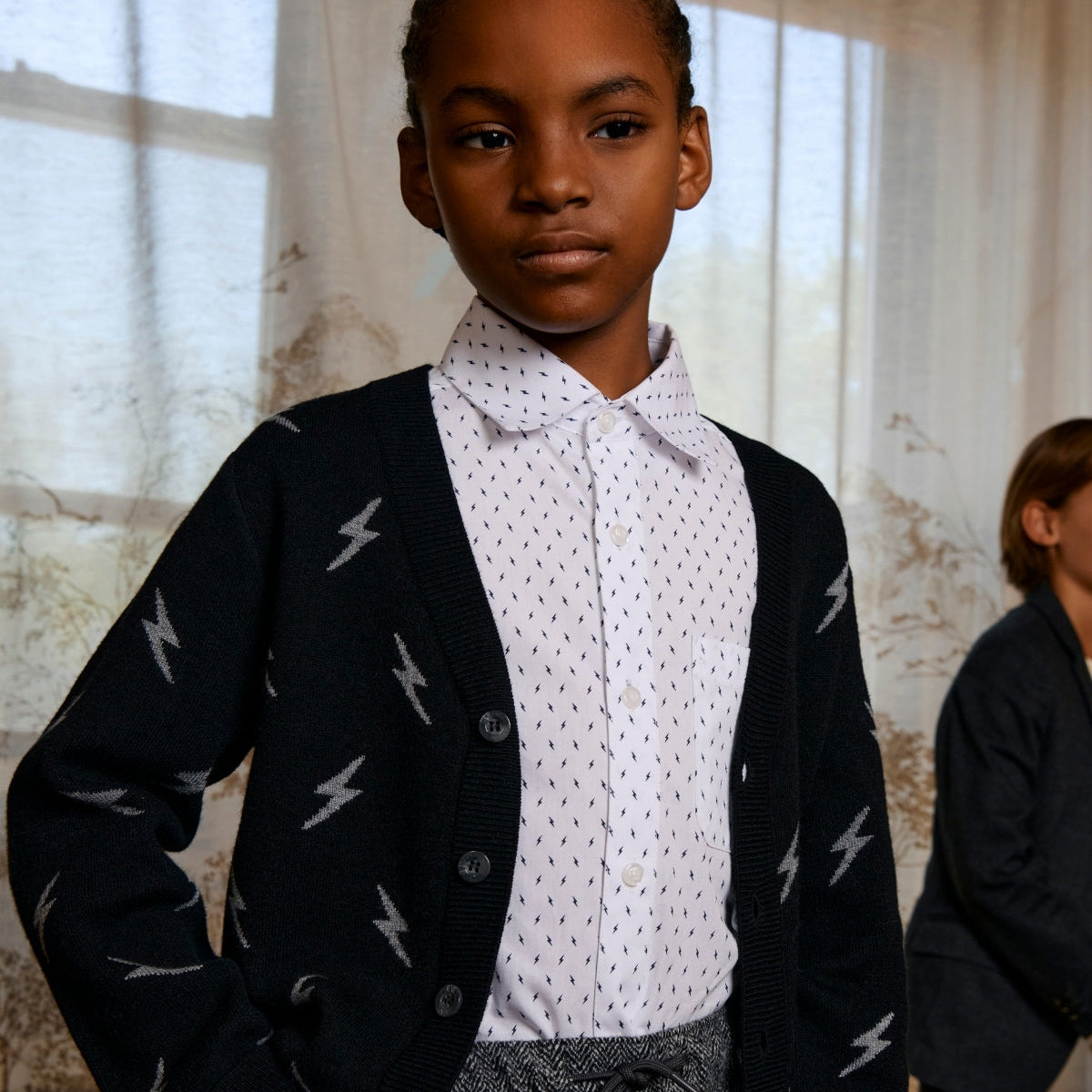 Young person wearing a black cardigan with white patterns, white shirt, and gray pants in a softly lit room.