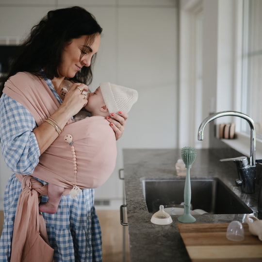 Woman holding a baby in a sling in a kitchen.