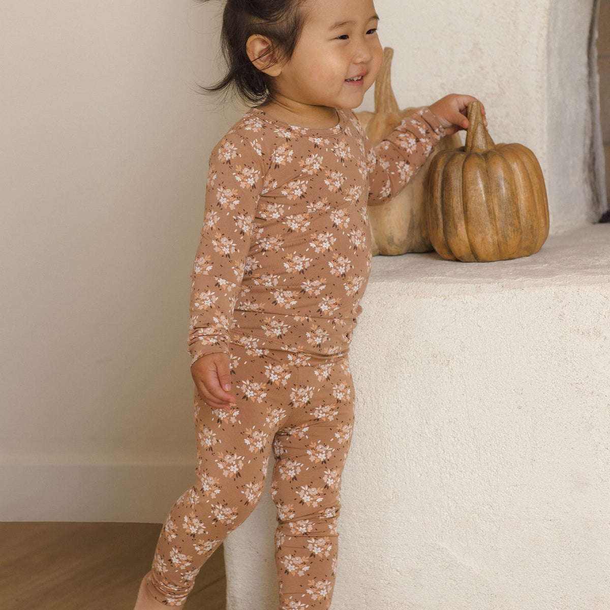 A toddler stands barefoot indoors next to a white textured wall, wearing a long-sleeve and matching pants pajama set in a warm tan color with a small white floral pattern. The child is touching a decorative pumpkin on a shelf.