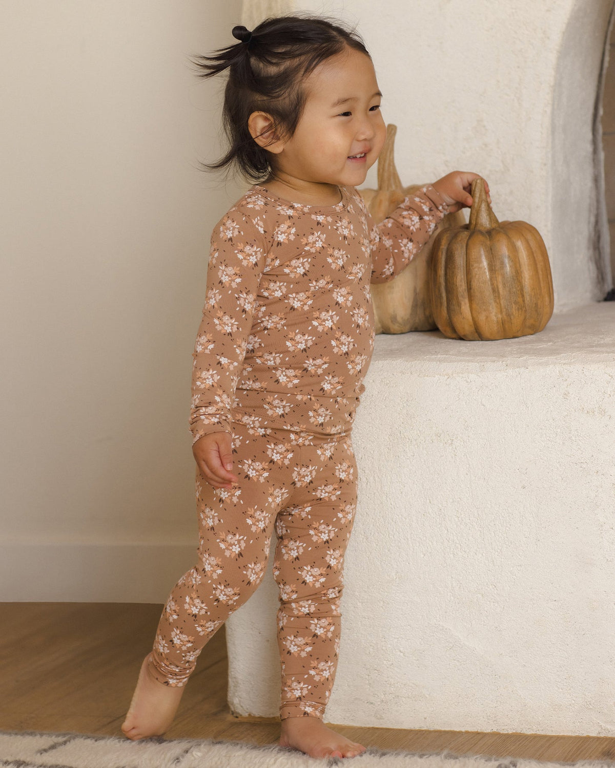 A toddler stands barefoot indoors next to a white textured wall, wearing a long-sleeve and matching pants pajama set in a warm tan color with a small white floral pattern. The child is touching a decorative pumpkin on a shelf.