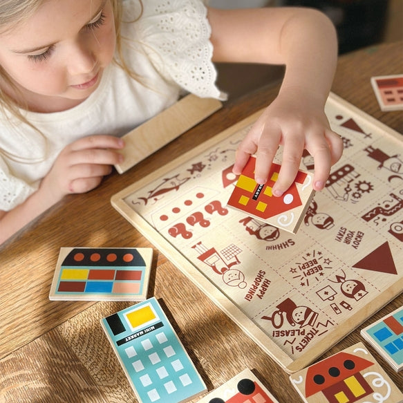 A young girl with blonde hair tied back sits at a wooden table, smiling as she plays with the Mentari Wooden Town Puzzle, holding a puzzle piece in her hand while assembling the colorful town scene.