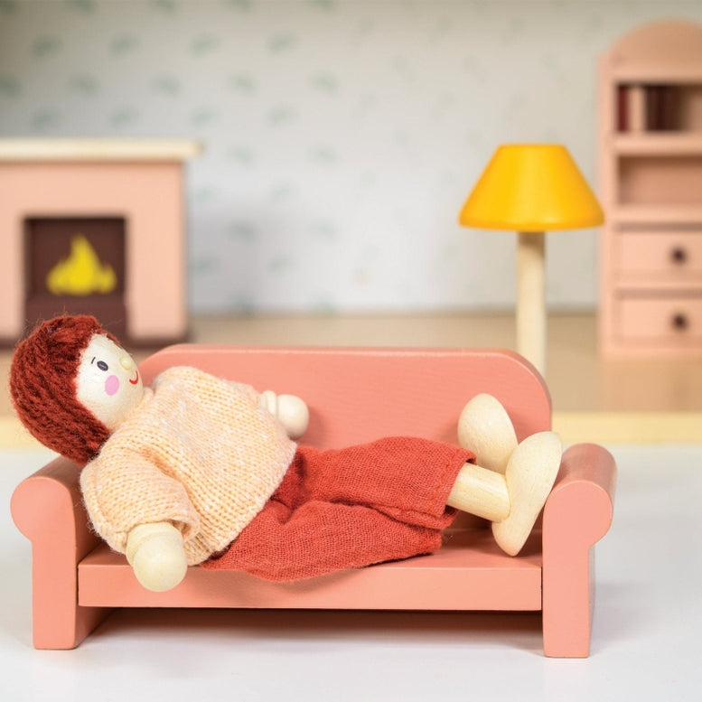 A close-up shot of a doll resting on a coral-pink toy sofa, wrapped in a knitted blanket, with a miniature wooden fireplace, a yellow floor lamp, and a bookcase in the background.