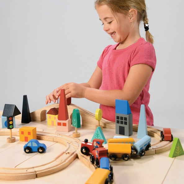 A young child with blonde hair tied back, wearing a pink short-sleeve shirt, reaches over a wooden toy train set laid out on a light-colored surface to place a red toy car onto the tracks. The setup features a figure-eight track configuration with a blue train engine and cargo cars, alongside colorful wooden town elements, including houses, trees, traffic lights, and a bridge.