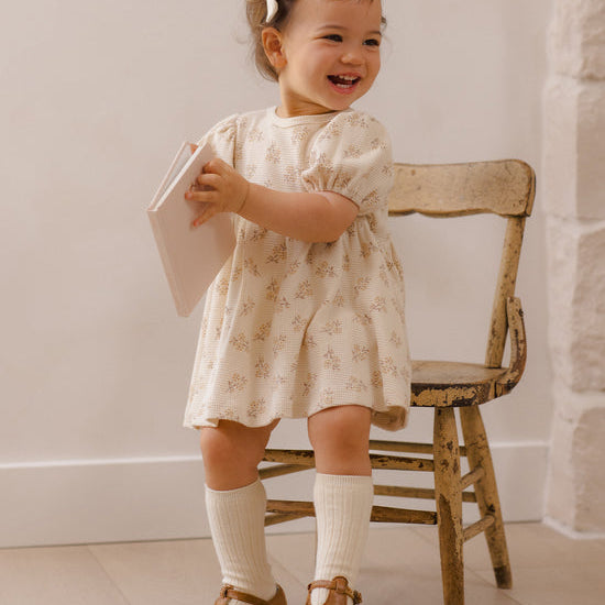 A toddler girl wearing a cream-colored short-sleeve dress with a delicate floral pattern, standing next to a rustic wooden chair and holding a book.