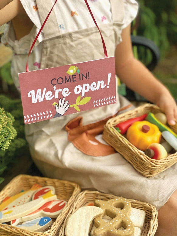 Person wearing an apron with a 'Come in! We're open!' sign, surrounded by toy food items.